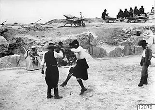 Men dancing on the orchestra of the theatre at Soli, with onlookers and wheelbarrows in the excavated cutting behind.