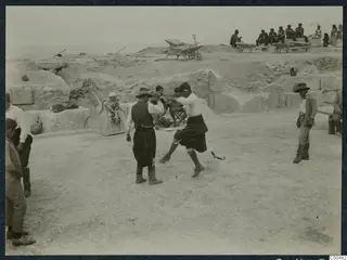 Men dancing on the stony floor of the theatre at Soli, with people sitting on wheelbarrows on the excavated ground behind.