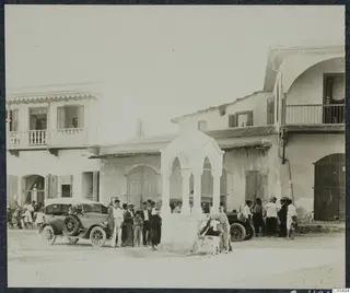 Square at Polis with a stone well canopy, two parked cars, and a crowd of men, a boy and a woman.