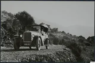 Open-top touring car with a registration plate driving along a gravel road in hills.