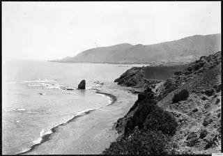 Rocky coastline with a sea stack and a curving pebble beach, with mountains rising behind in the Kyrenia Mountains