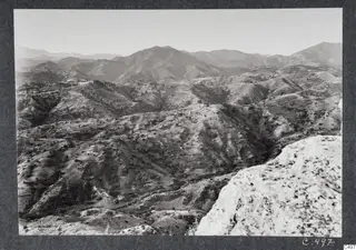 Wide view over rounded hills and valleys, with distant mountains under a hazy sky and a pale rock in the foreground.
