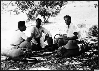 Three men eating outdoors on the ground, seated on sacks, with a pith helmet beside the man on the right.