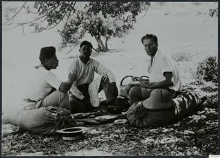 Three men eating outdoors, seated on sacks under a tree, with a pith helmet resting beside one of them.