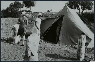 Two women and a man beside a tent at Vouni, with three stone sculptures standing outside it.