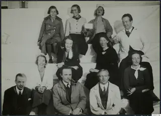 Group of eleven people posed in three rows on white steps, with a girl at top left and a man with a pipe in the front row.