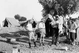 Expedition camp at Vouni with three tents, and people standing around a tree from which an animal hangs for slaughter
