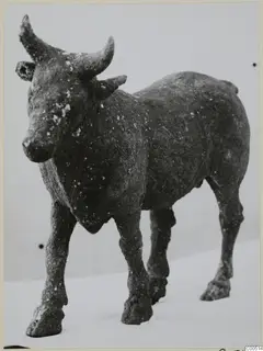 Bronze figure of a standing cow or bull with curved horns and a corroded dark surface, shown against a pale background.