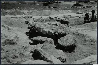 Excavation at the Temple of Athena, Vouni, with fallen stone blocks in a trench and two people seated at the right edge