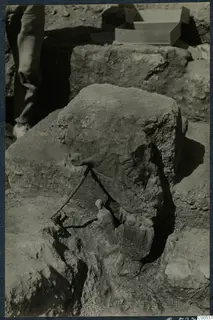 Small standing figurine and a cylindrical object in excavated ground at Vouni Palace, with a large stone block above them