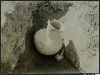 Large pale jug and a small dark vessel lying in an excavated trench at Vouni