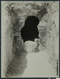 Narrow excavated passage leading to a dark arched opening, with a large pale stone block lying in front of it