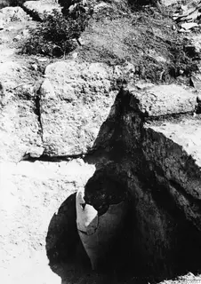 Broken pottery vessel standing in soil during excavation at Vouni Palace, with pale stone blocks above it.