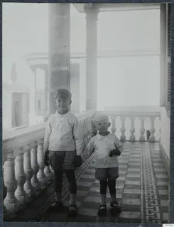 Two young children standing on a tiled veranda beside a stone balustrade and tall columns.