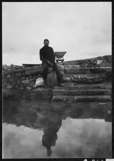 A man sits on stone steps and a stone wall at Vouni, with a pool of water reflecting him in the foreground.