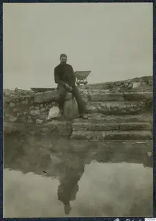 Man seated on a stone block beside stone steps and walls at Vouni, with still water in the foreground reflecting him