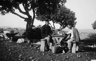 Two men sit at a wooden table under a tree at Vouni, one of them eating.