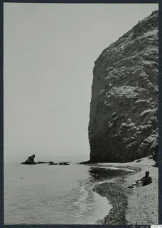 The bathing beach at Vouni, with a tall rock cliff on the right and a seated person on the pebbles.