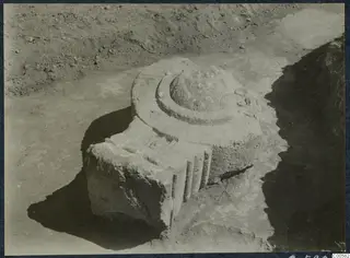 Carved stone architectural piece lying in an excavated trench at Vouni Palace, with a stepped round top and vertical grooves