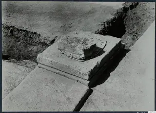 Squared stone block with a broken stone fragment on top, among slabs in an excavation at Vouni