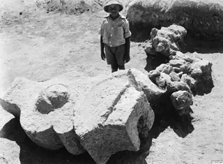 Child standing behind a large carved stone block at Vouni, with broken stones and a trench wall beside it