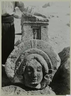 Limestone head carved in relief on a slab at Vouni, with a niched stone block behind it and a person at the left edge