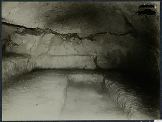 Interior of a rock-cut tomb chamber at Vouni with carved benches and a sunken rectangular area in the floor.