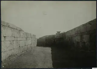 Excavated passage between two high ashlar walls at Vouni palace, with a stone basin on top of a wall in the distance.