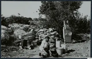 Eleni crouching outdoors to wash dishes beside a stone wall, with plates drying on a wooden rack behind her.