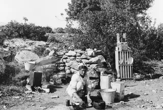 Eleni kneeling outdoors at Vouni, washing dishes beside a stone wall, with a dish rack and buckets nearby.