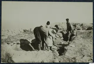 Workers bending over an excavation trench at Vouni, with a rail track along the edge.