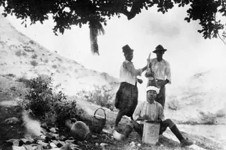 Three men at Vouni under a tree: one seated with a tin, two standing behind him, one holding a hoe over his shoulder.