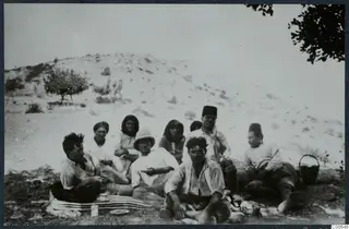 Nine people sit and rest on a striped mat by a hill at Vouni, with cups and a basket nearby.