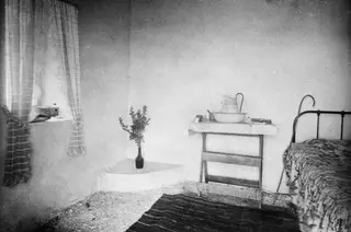 Interior of a whitewashed room at Vouni with a curtained window, a washbasin on a table and an iron bed.