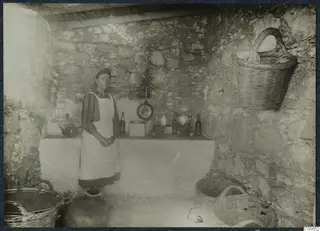 Eleni standing in a stone-walled kitchen at Vouni, with a frying pan, pots, bottles and baskets