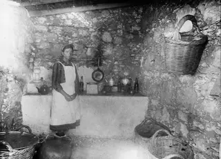 Eleni standing in a stone-walled kitchen at Vouni, with a frying pan, pots, a teapot and baskets