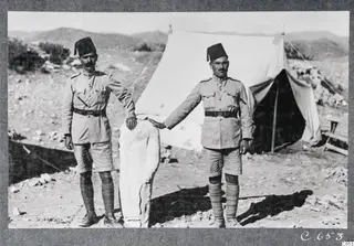 Two men in uniform and fez beside a pale limestone statue, in front of a tent at Vouni