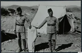 Two men in khaki uniform and fez caps beside a pale limestone statue, with a white tent behind them at Vouni