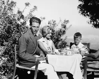 Three people at breakfast at a laid round table outdoors, with bushes behind them.