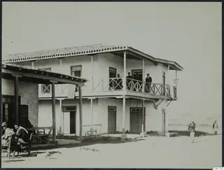 Two-storey whitewashed building with a timber-railed balcony, men standing on it and seated men in wooden chairs outside