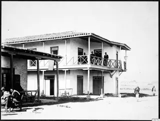 Two-storey whitewashed building with a wooden balcony, men standing on it and seated on chairs under the porch