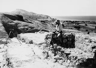 Alfred Westholm sits on a stone wall at the excavation at Soli, with hills and the sea behind him.
