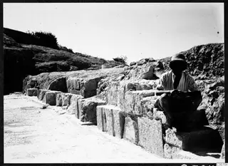 Large cut stone blocks forming a wall face at Soli, with a seated man in a wide-brimmed hat on the right.