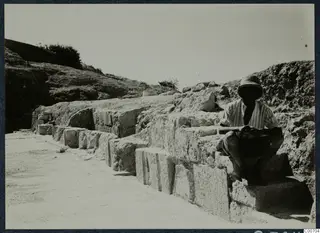 Seated man in a striped shirt and hat on cut stone blocks of the stage facade at Soli theatre