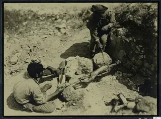 Two men roasting meat on a spit over stones at Soli