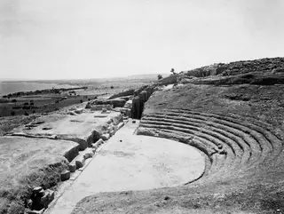 Excavated theatre at Soli with curved stone seating rows around a paved orchestra, with the coast and fields beyond.