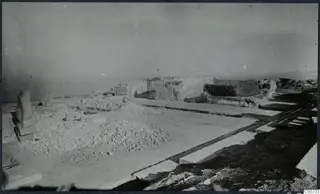 Excavated area at Vouni palace with rubble heaps, an upright stone, long stone slabs in the foreground and ruined walls behind.