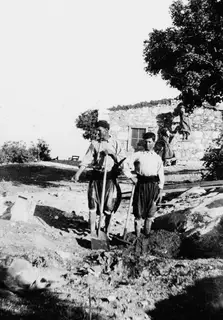Two men leaning on spades beside a stone house under construction at Vouni