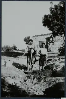 Two men leaning on spades beside a stone house under construction at Vouni