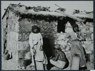 Two women and a crouching man beside a stone house with tiles and timbers on its roof at Vouni